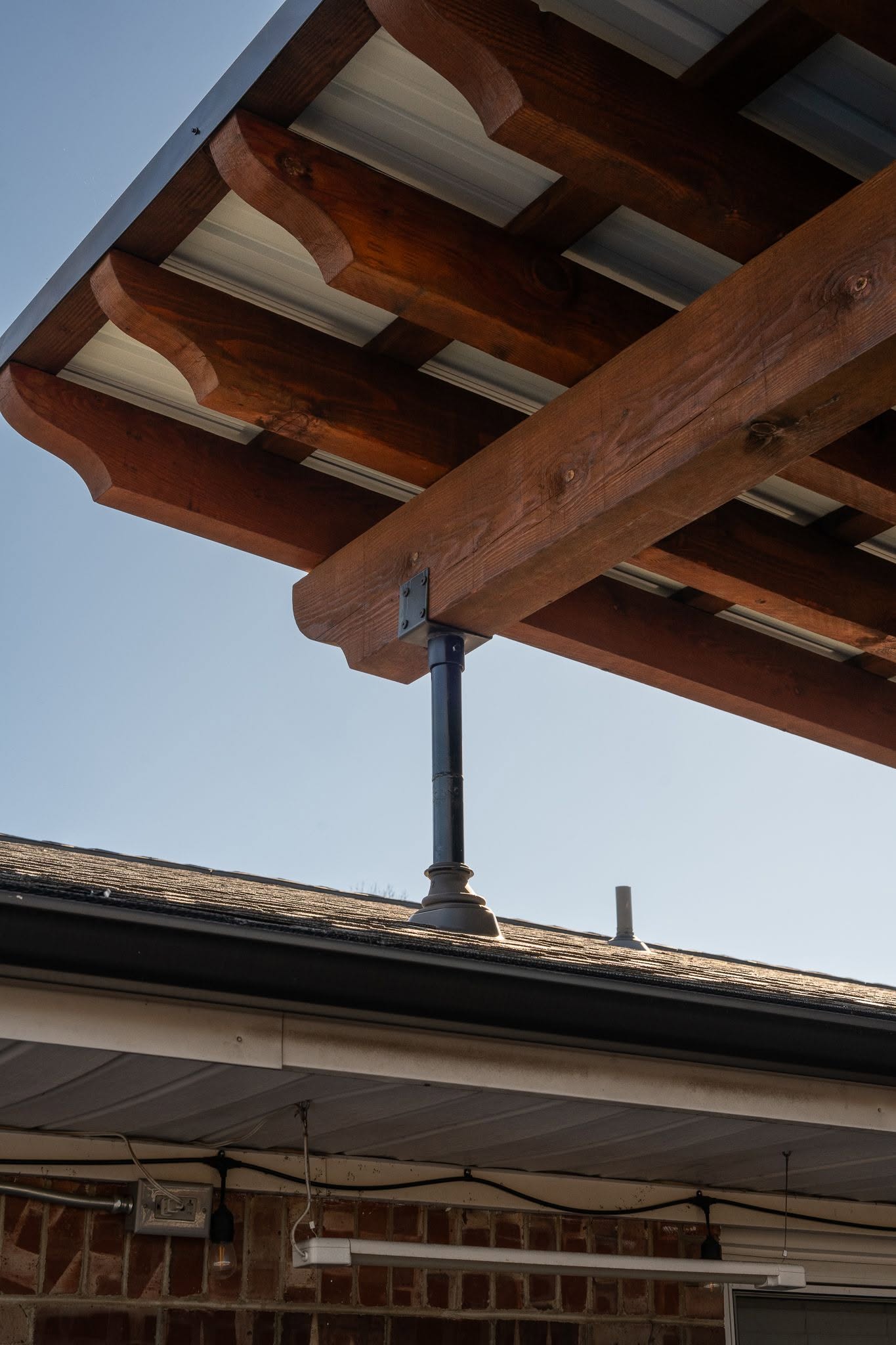 A wooden pergola with a metal roof is supported by a dark pole over a house.