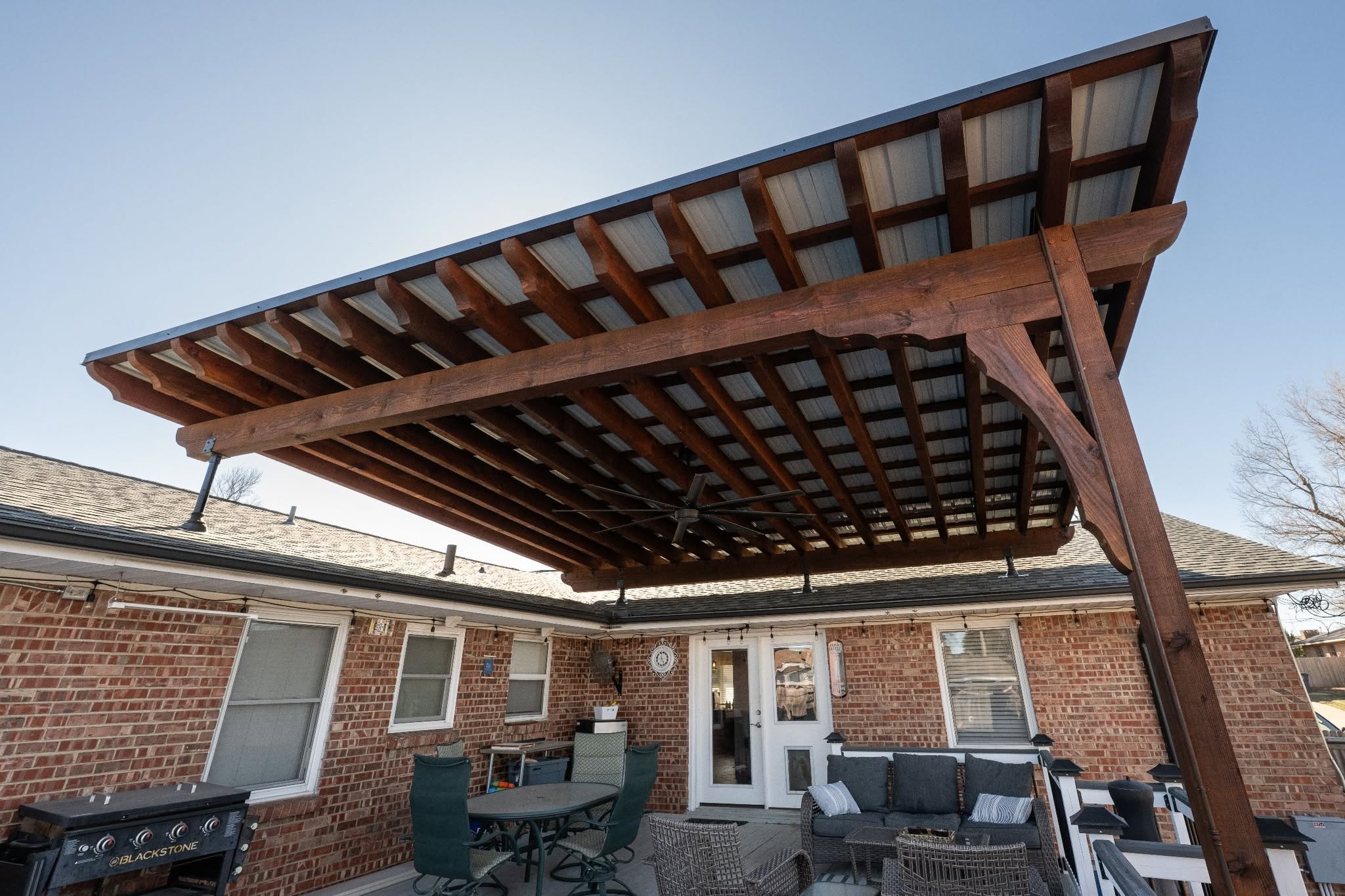 A covered patio with a wooden pergola, metal roof, and outdoor furniture is shown on a sunny day.