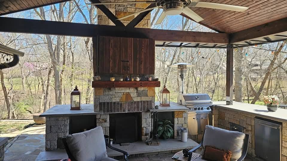 An outdoor kitchen with a grill, fireplace, and seating is set against a wooded backdrop.